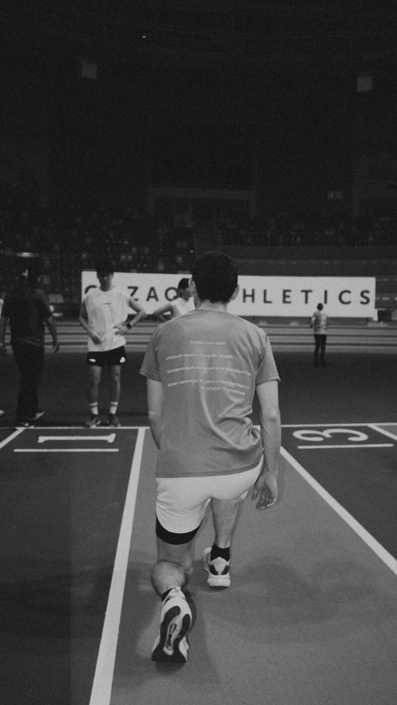 A grayscale shot shows a man in athletic wear walking on a track with the word "ATHLETICS" visible in the background. The man's shirt features text that reads "MARATHON team" along with other smaller lines of text in a foreign language.