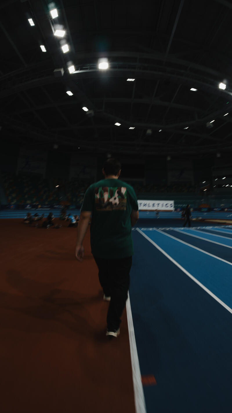 A person in a green t-shirt walks along a blue and red track in a stadium; a sign with the word "ATHLETICS" is visible.