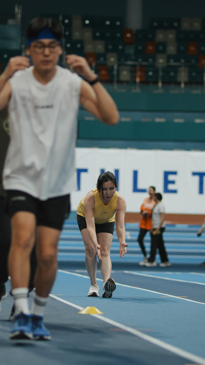 A woman in a yellow tank top and black shorts is stretching her leg on a blue running track, with blurred figures of other people in the background and the text "TLET" partially visible.