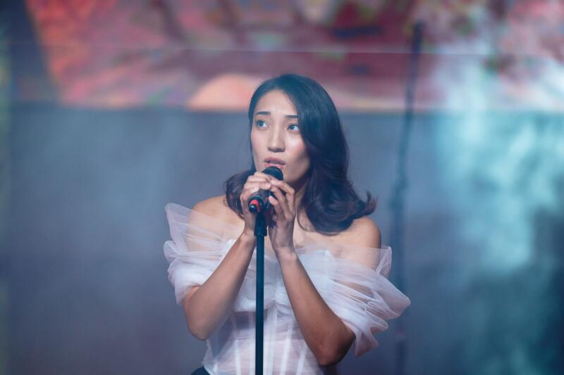 A woman with dark hair is singing into a microphone while wearing a white, sheer, off-the-shoulder top.