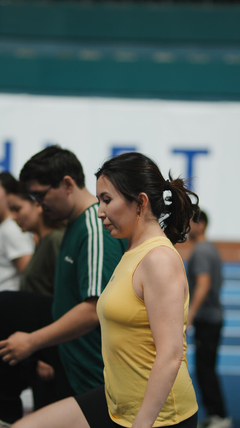 A woman in a yellow tank top stands in a line with other people; a man with glasses wears a green shirt with white stripes.