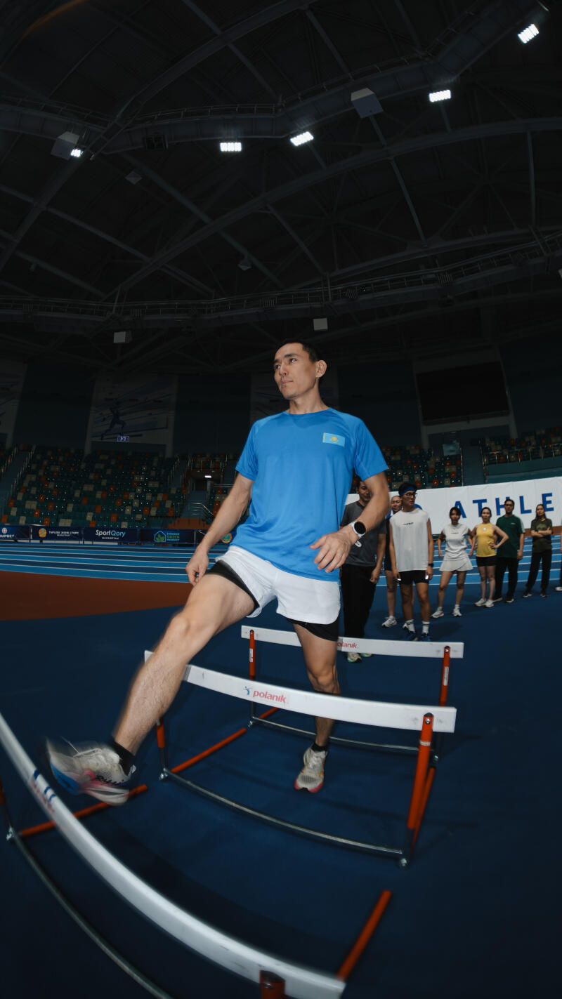 In an indoor stadium, a man in a blue t-shirt and white shorts is jumping over hurdles, with a group of people standing in the background.