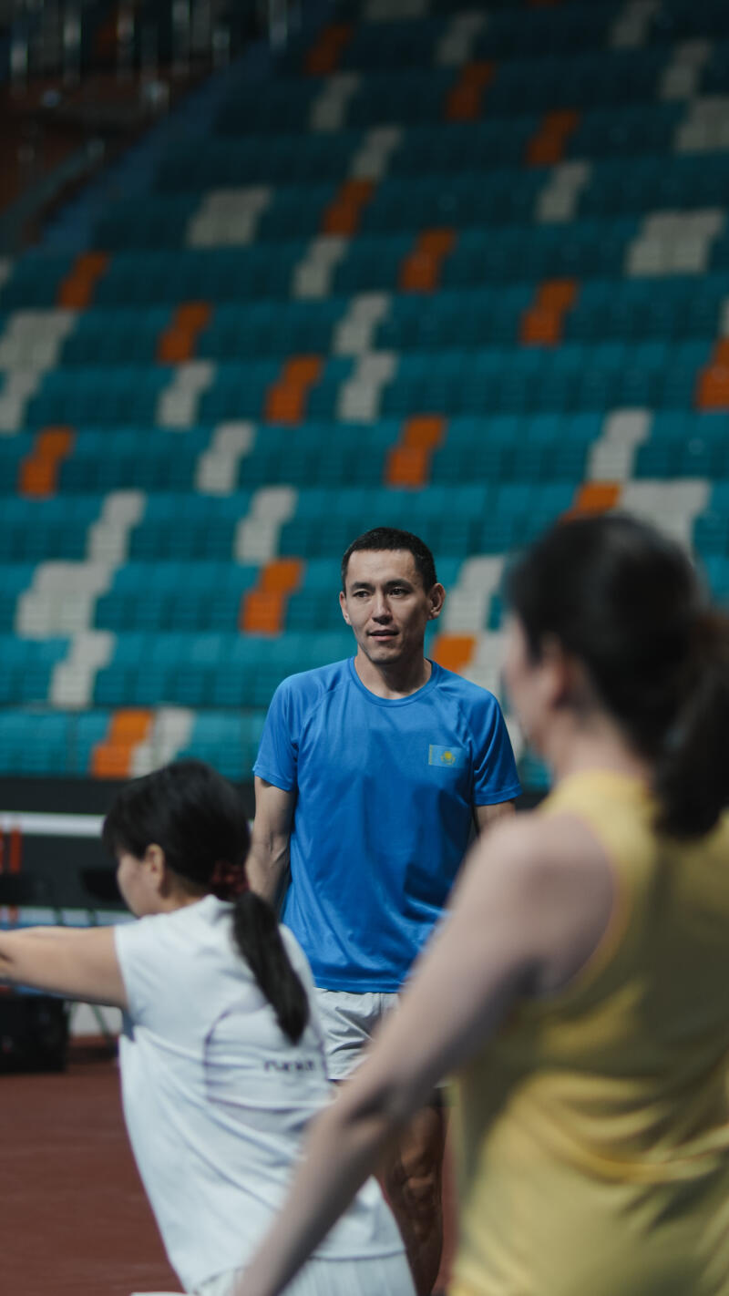 The image shows a man in a blue shirt with the Kazakhstan flag on it, standing on a track in front of stadium seats, with two women in the foreground.
