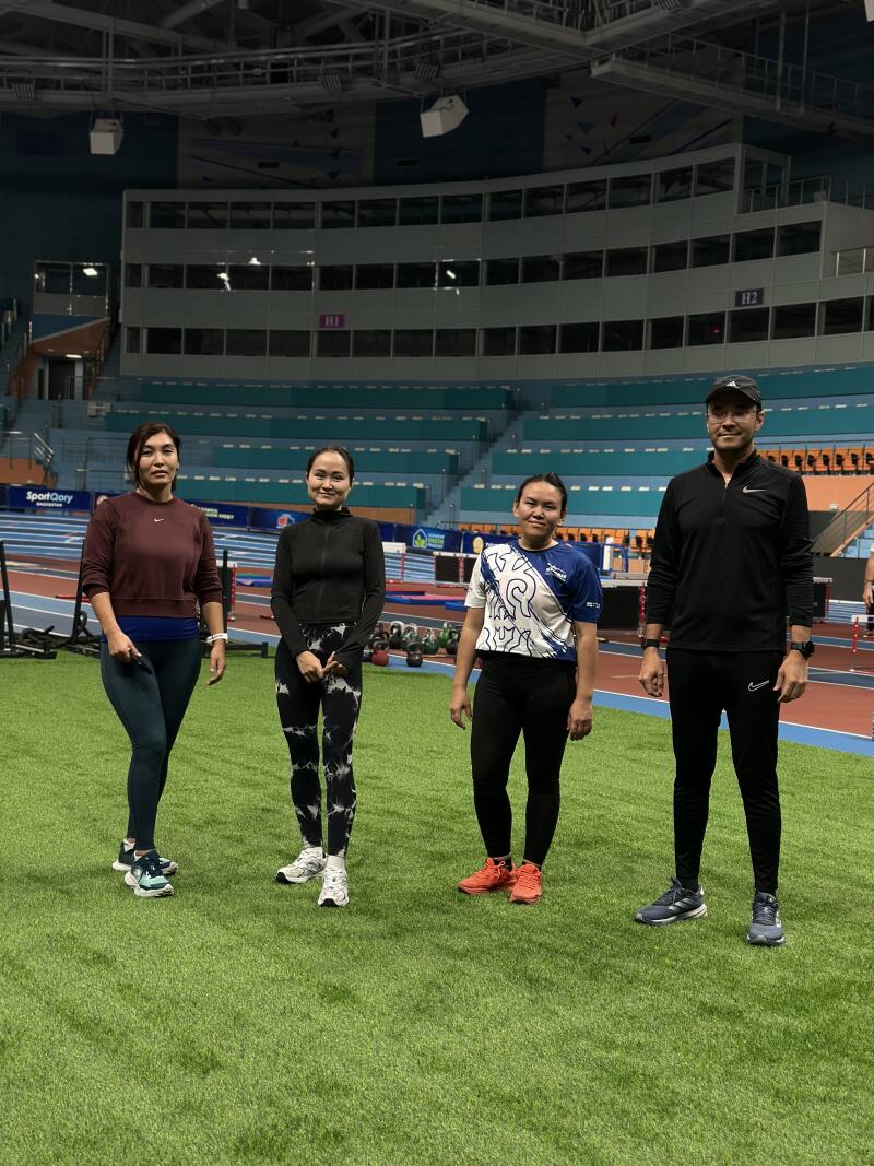 Four people stand on an indoor green field with stadium seating in the background, including three women wearing athletic attire and one man in a black athletic suit and baseball cap.