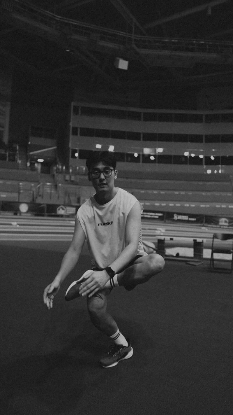 In a black and white shot, a person wearing glasses, a tank top with the word "runk!" on it, shorts, socks, and sneakers is squatting with one leg resting on the other knee.