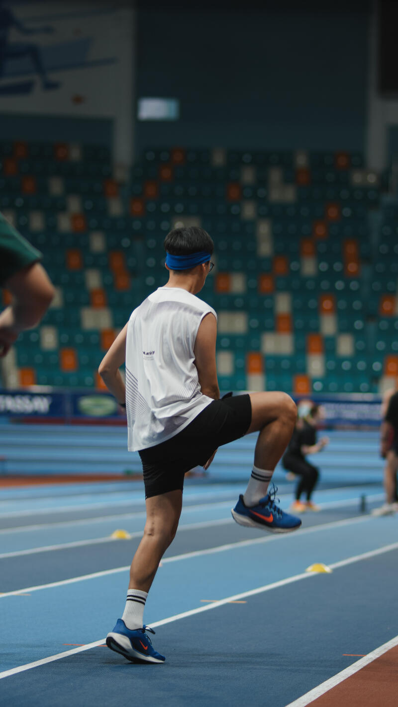 A person wearing a white tank top, black shorts, blue headband, and glasses is doing a high knee exercise on a blue indoor track, with stadium seating in the background.