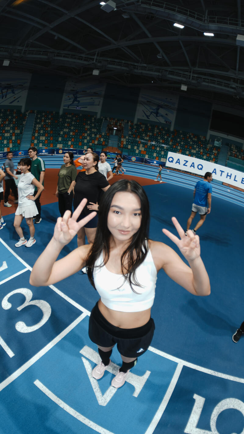 A young woman in athletic wear flashes a peace sign while standing on a blue running track with lane numbers, with other people and the words "QAZAQ ATHLE" visible in the background.