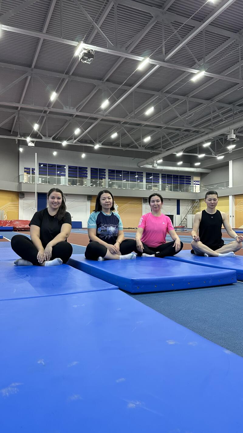 Four women are sitting cross-legged on blue mats in a large, brightly lit gymnasium.