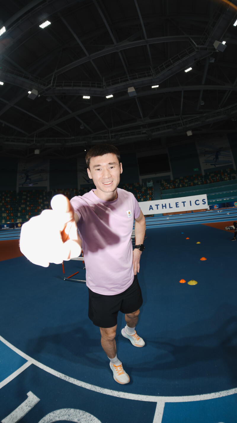 A man in sportswear points toward the camera in an indoor athletic facility, with an 'ATHLETICS' sign visible in the background and colorful cones on the blue track.