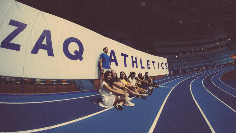 A group of people are sitting and standing in front of a large banner that reads "ZAQ Athletics" on a blue indoor track.