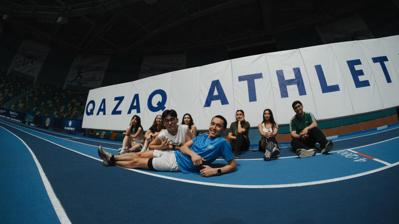 A group of people are sitting on a blue athletic track in front of a banner that reads "QAZAQ ATHLETICS".
