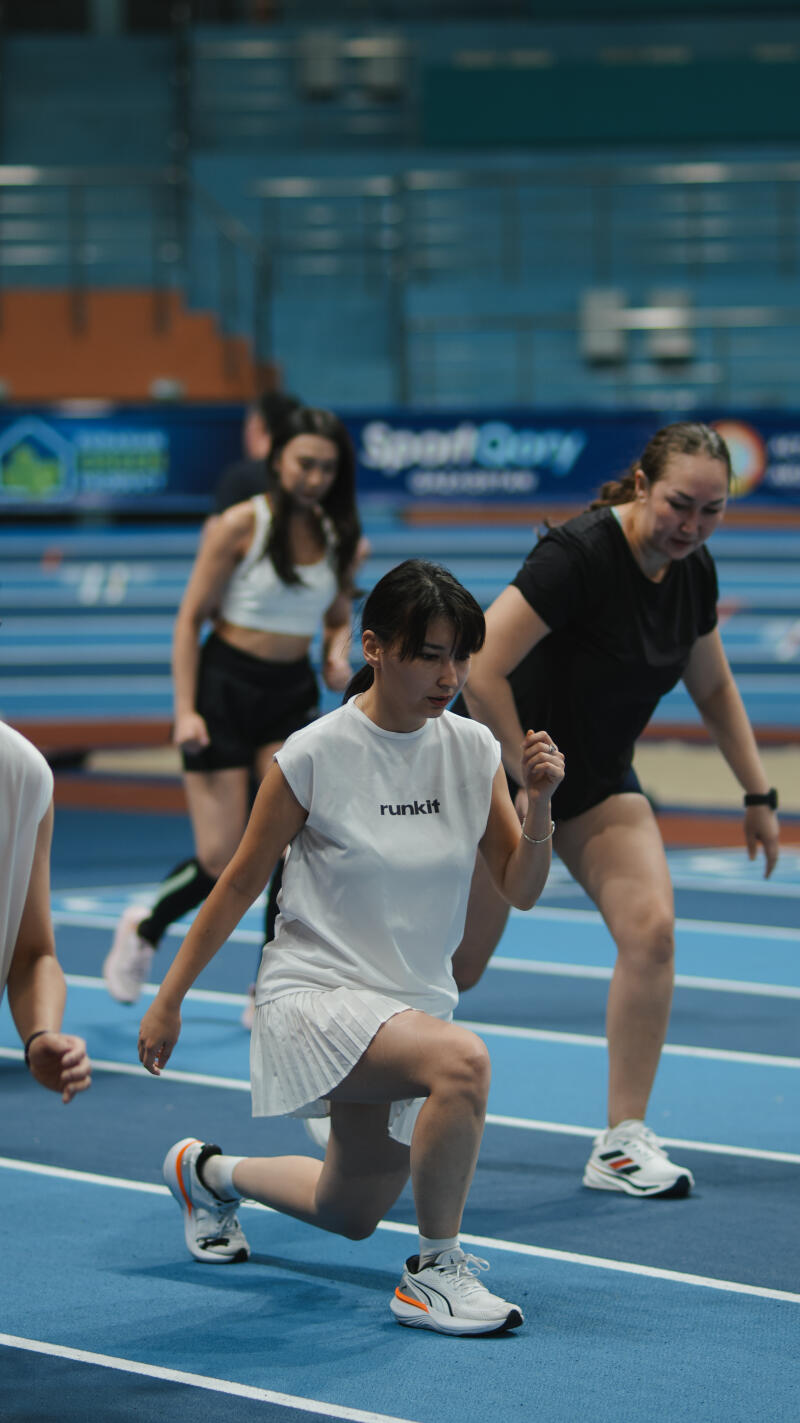 A group of women are exercising on a blue track, with the woman in the foreground wearing a white 'runkit' t-shirt and a white pleated skirt, performing a lunge.