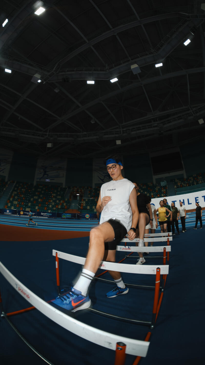 A man in a white tank top and blue headband steps over hurdles on a blue track, with other people visible behind him.
