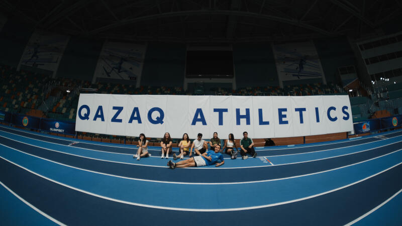 A group of people are sitting and lying on the blue running track of an indoor stadium under a banner that reads "QAZAQ ATHLETICS".