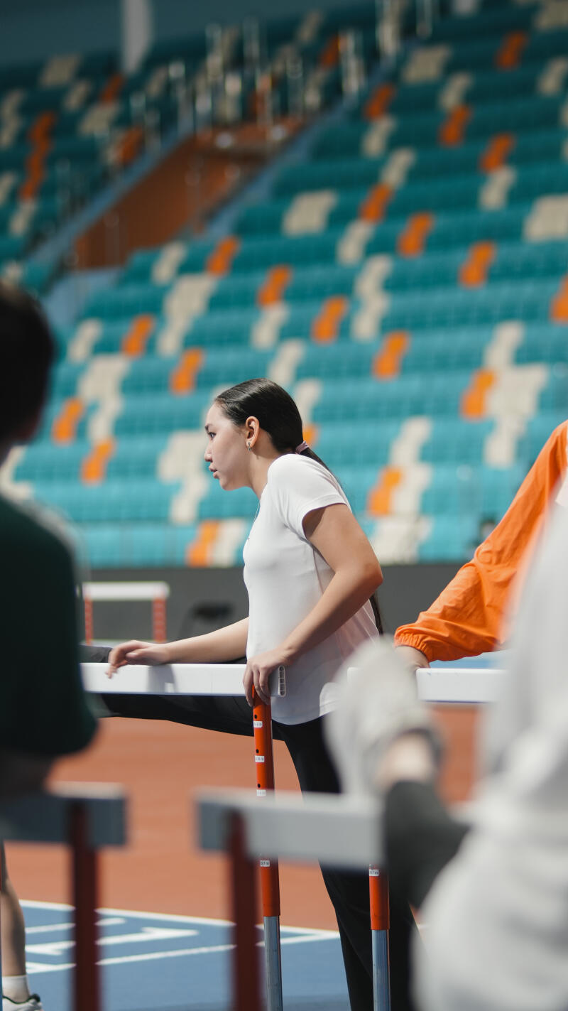 A woman in a white shirt stretches her leg over a hurdle on a track, with blue and orange stadium seats in the background.