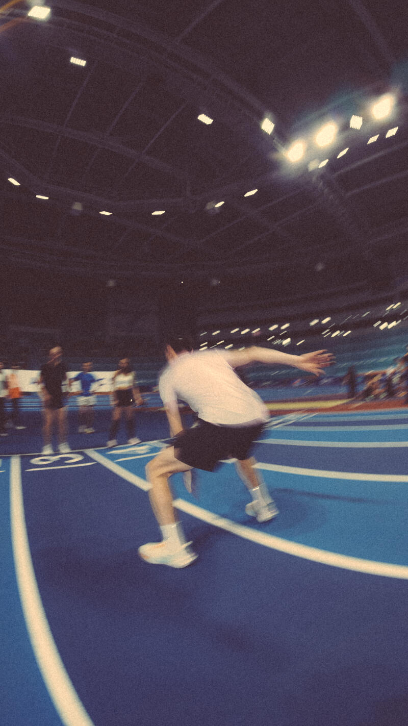 The image shows a person in motion on a blue track with white lines, wearing a white t-shirt, black shorts, white socks, and white shoes, with other people visible in the background and bright lights overhead.