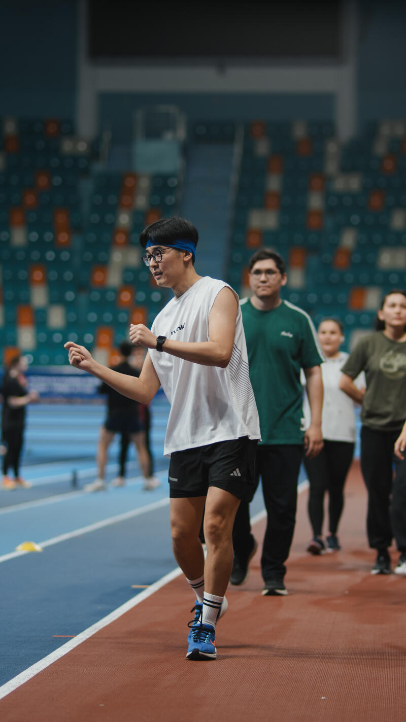 A man in a white tank top and black shorts walks on a track with other people in the background; he is wearing a blue headband and glasses.