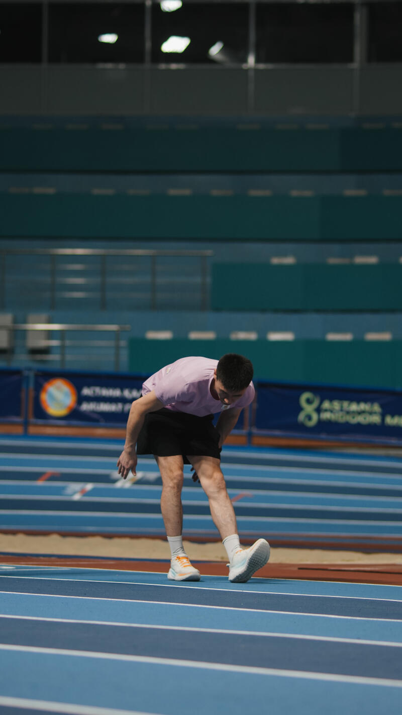 A man in a pink shirt and black shorts stretches his leg on a blue indoor track; the text "ASTANA INDOOR MET" is visible in the background.