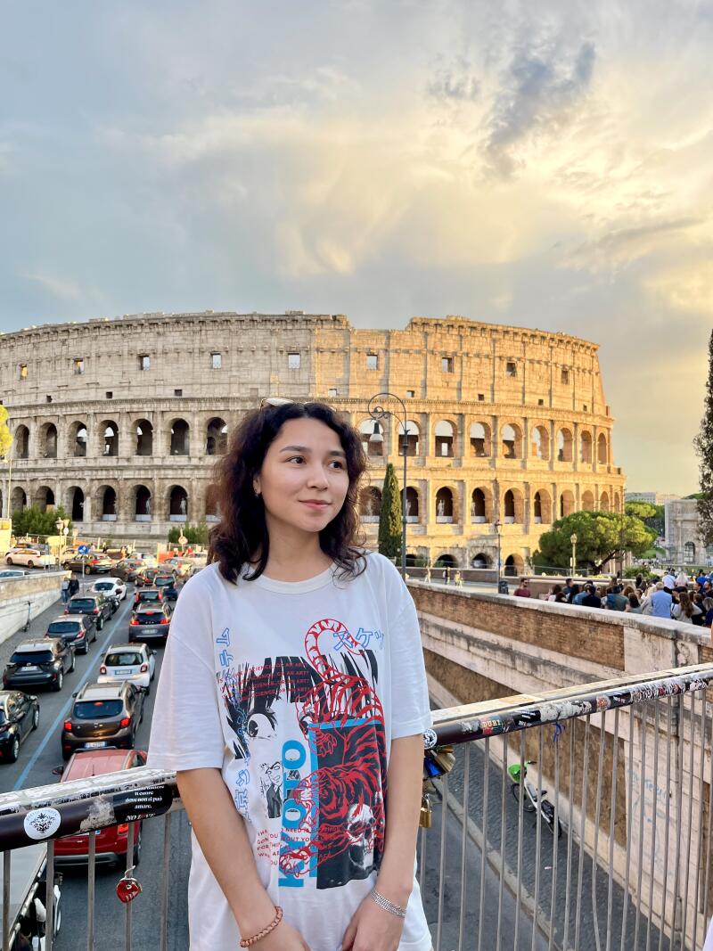 A young woman in a graphic t-shirt stands on a bridge with the Colosseum in the background.