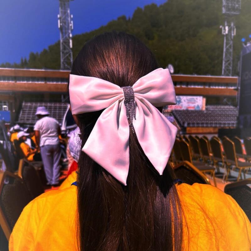 A person with long dark hair wears a large pink bow with a sparkly center at the back of their head; in the background, there are seats and a tower structure.