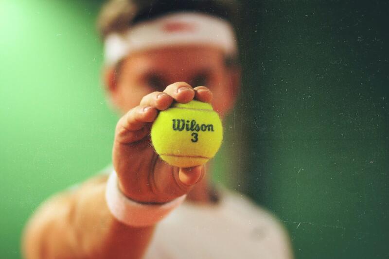 A person is holding a tennis ball with the text "Wilson 3" printed on it, wearing a white headband and wristband.