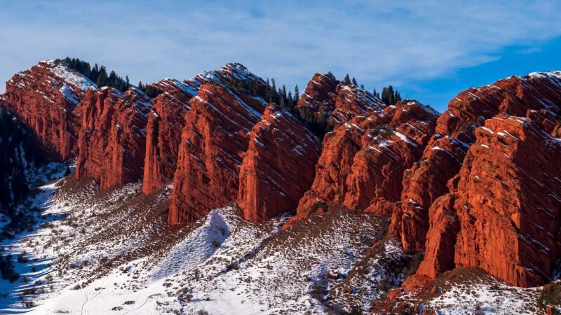 The image shows a mountain range with red rock formations partially covered in snow, set against a blue sky.