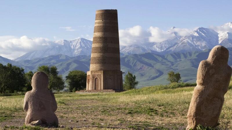 The image shows the Burana Tower and two balbals on a grassy field, with a backdrop of mountains and a blue sky.