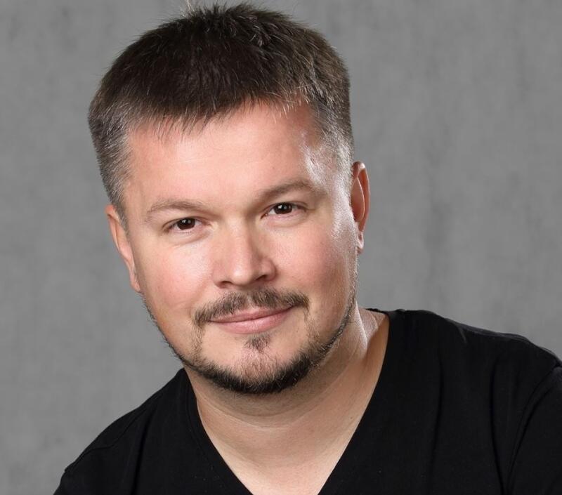 A portrait of a man with short brown hair, a neatly trimmed beard and mustache, wearing a black V-neck shirt against a gray background.