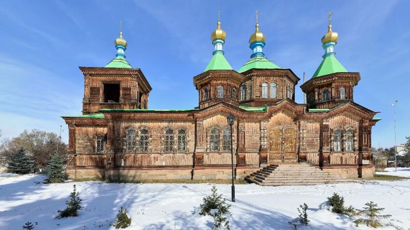 A wooden church with green roofs and golden domes stands in a snowy landscape under a blue sky.