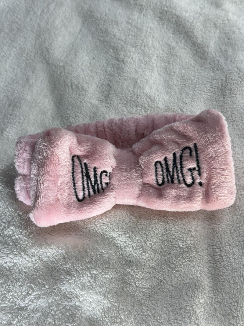 A pink plush headband with the text "OMG!" embroidered in black on both sides, lying on a white fuzzy surface.