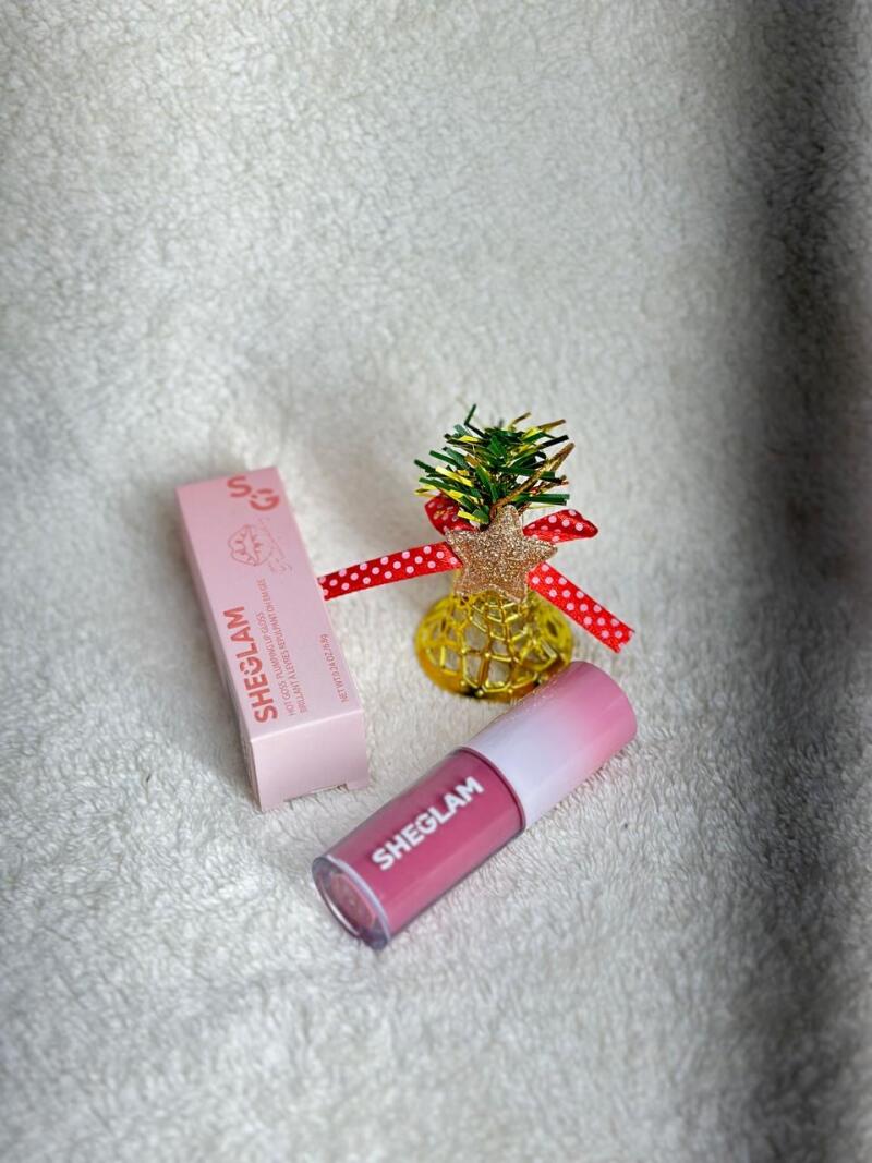The image shows a tube of SHEGLAM lip gloss and its packaging box, alongside a decorative Christmas tree ornament with a red polka dot ribbon, all on a white textured surface. The box reads: SHEGLAM, HOT GOSS PLUMPING LIP GLOSS, BRILLANT A LEVRES REPULPANTOH EM GEE, NET WT 0.24 OZ/6.8g.