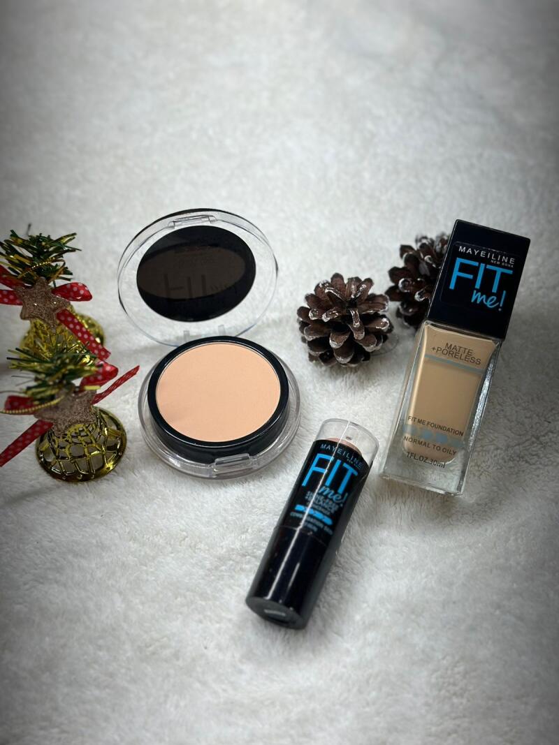 The image shows several makeup products from Maybelline's "Fit Me!" line, including foundation, powder, and a stick concealer, arranged with pine cones and a small Christmas tree on a white, textured surface.