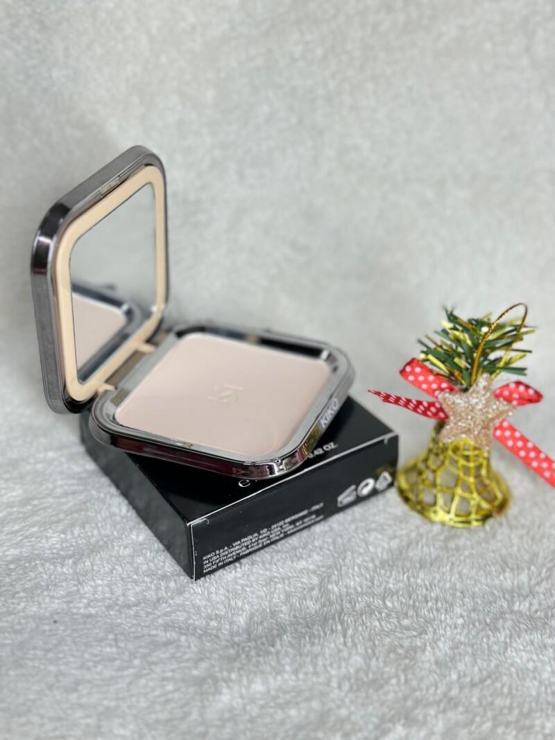 The image shows an open Kiko Milano compact powder case with a mirror in the lid, sitting on its black packaging next to a decorative bell.