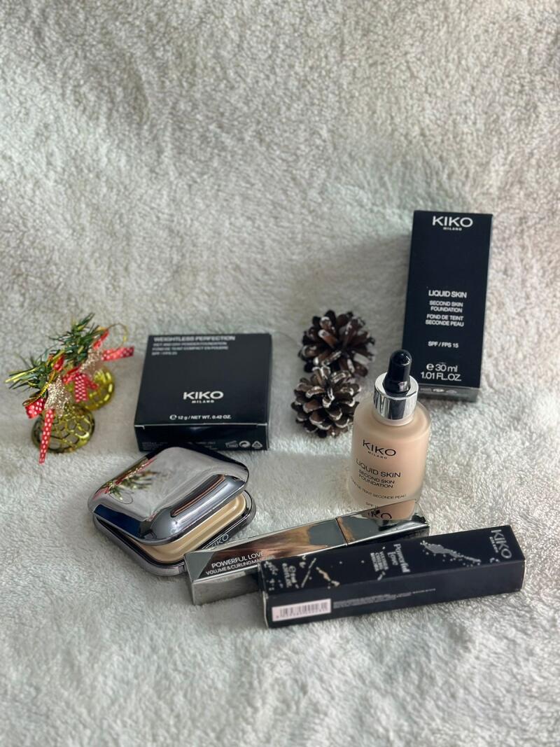 The image shows an assortment of Kiko Milano makeup products, including liquid skin foundation, weightless perfection powder, and powerful love volume & curling mascara, arranged with pine cones and Christmas decorations on a white textured surface.