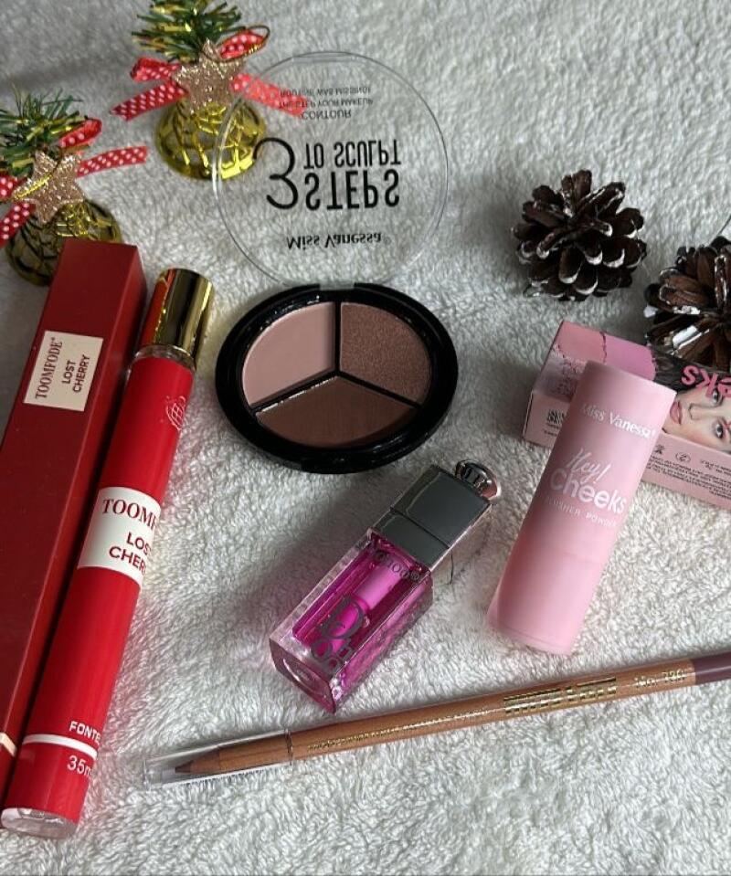 The image displays various makeup products, including eyeshadow palettes, lip gloss, lip liner, and blush, along with Christmas ornaments and pine cones on a white textured surface. Text visible includes 'TOOMFODE LOST CHERRY', '3 SECRETS', and 'Miss Vanessa Hey! Cheeks'.