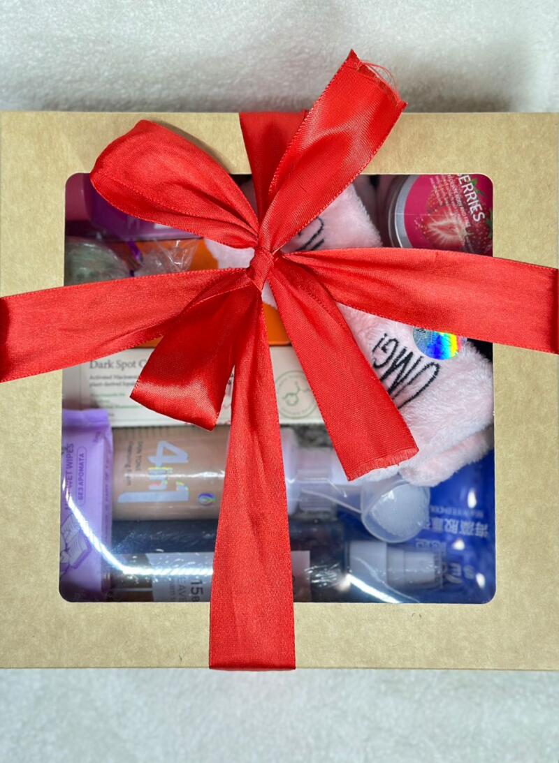 A gift box with a variety of beauty and skincare products is tied with a large red ribbon. The box includes items such as facial wipes (БЕЗ АРОМАТА WET WIPES), a dark spot corrector (Dark Spot C), and a product labeled 'OMG!'