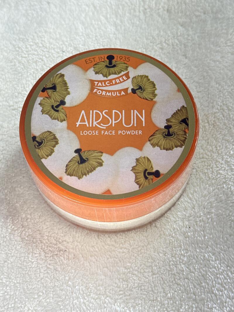 A container of Airspun Loose Face Powder sits on a white textured surface. The container is round with an orange lid, and the label reads "EST. IN 1935", "TALC-FREE FORMULA", and "AIRSPUN LOOSE FACE POWDER". The label also features a pattern of flower-like designs.