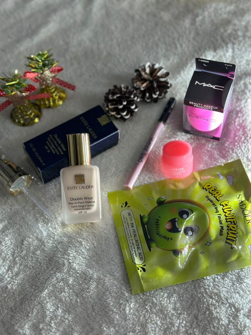 The image displays an assortment of beauty products, including Estee Lauder Double Wear foundation, a MAC beauty sponge, Laneige lip sleeping mask, Sadoer kiwi fruit facial mask, a pink eyeliner, pine cones, and decorative Christmas trees.