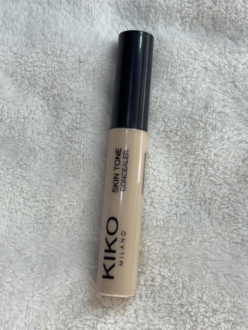 The image shows a tube of Kiko Milano Skin Tone Concealer with a black cap, set against a white, textured background.