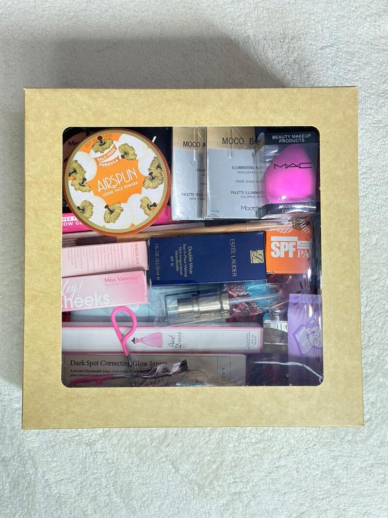 A collection of makeup products is visible through the transparent window of a beige cardboard box, including Airspun loose face powder, MOCO A and MOCO B palettes, a MAC makeup sponge, Estee Lauder Double Wear foundation, and Miss Vanessa blusher powder.
