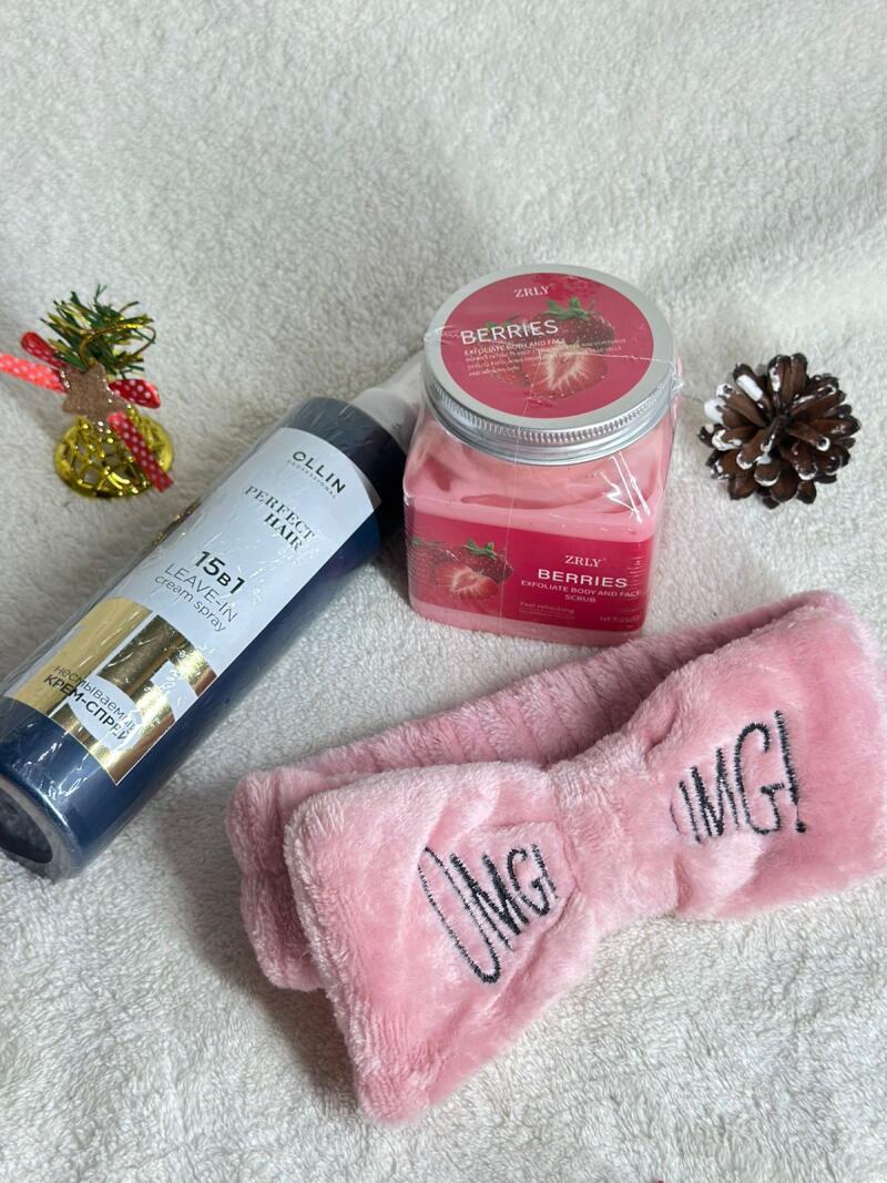 A flat lay shows Ollin Perfect Hair 15-in-1 Leave-in Cream Spray, Zrly Berries Exfoliate Body and Face Scrub, and a pink headband with the text "OMG!" on a white towel, along with a decorative pinecone and small Christmas ornament.