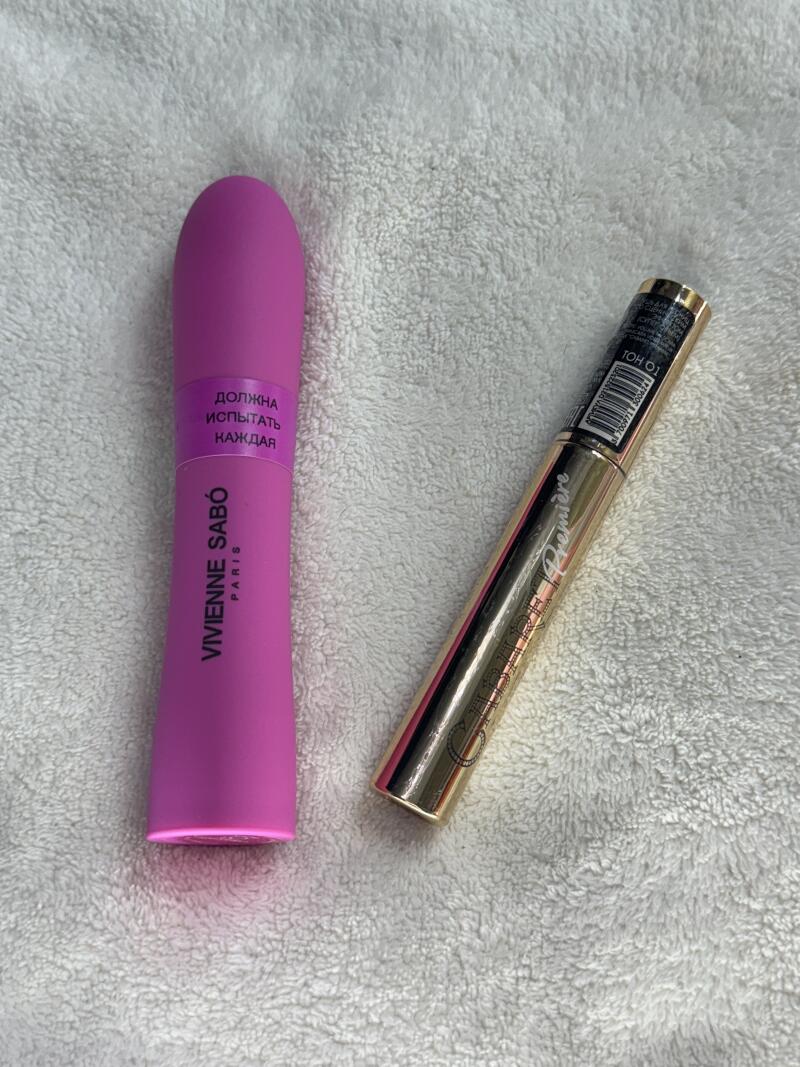 The photo shows two tubes of mascara on a white towel: one is pink and labeled "VIVIENNE SABÓ PARIS" with the text in Russian "ДОЛЖНА ИСПЫТАТЬ КАЖДАЯ", and the other is gold and labeled "CLARINS Premiere".