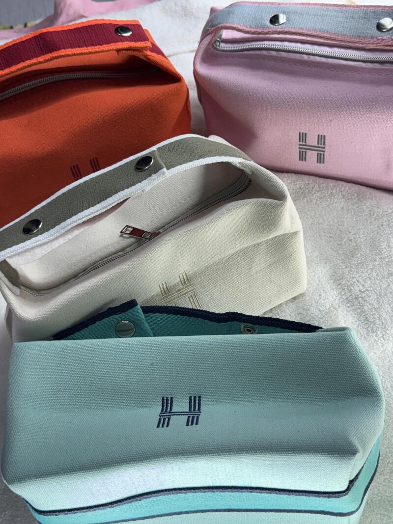 The image shows four cosmetic bags in different colors: orange, pink, beige, and light blue, each featuring a distinctive 'H' logo. They are made of canvas with zipper closures and snap details.