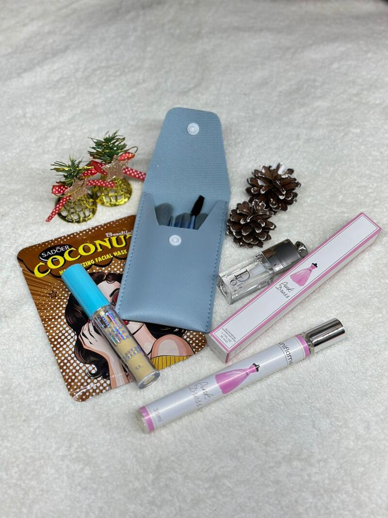 The image shows a collection of beauty products including a Sadoer Coconut facial mask, a makeup brush set in a light blue case, a tube of concealer with a blue cap, a bottle of Dior perfume, and a Uniflame Pink Dress perfume stick. There are also two small pine cones and two tiny decorative Christmas trees in the shot, all arranged on a white, textured surface.