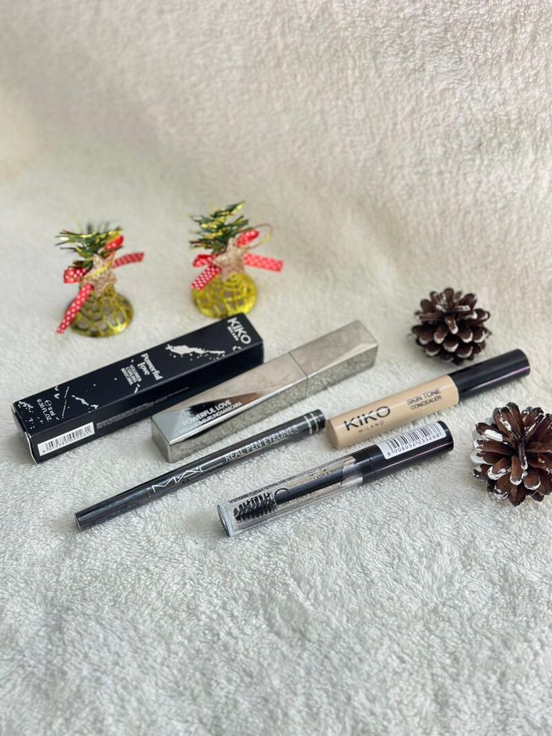 The image shows a collection of makeup products including Kiko Milano mascara and concealer, a MAC eyeliner, and a clear brow gel, styled with two miniature Christmas trees and pine cones on a white textured surface.