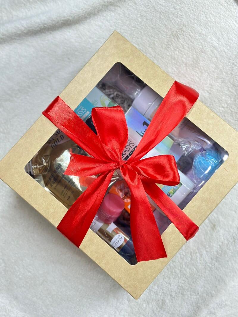 A square, light-brown box with a transparent window is filled with various beauty products and tied with a bright red ribbon.