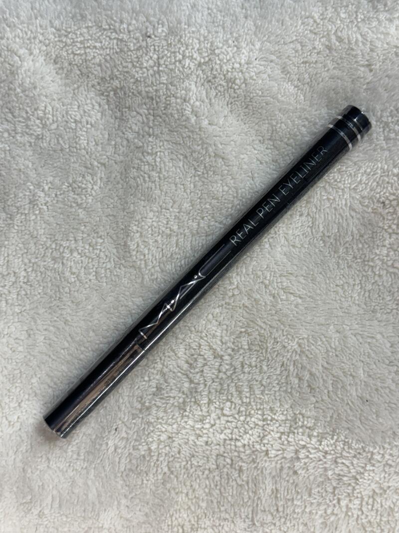 A black MAC 'Real Pen Eyeliner' is diagonally placed on a white, textured surface.