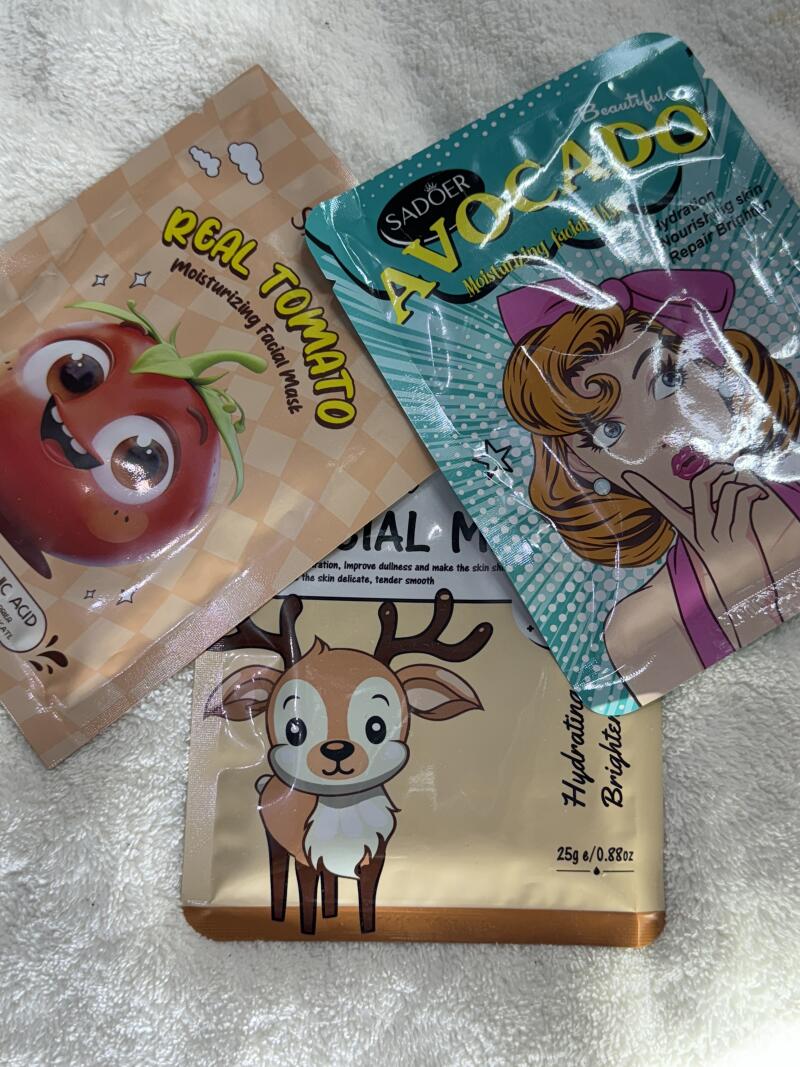 The image shows three facial mask packets with different designs: a cartoon tomato labeled 'REAL TOMATO Moisturizing Facial Mask', a retro cartoon woman with the text 'SADOER AVOCADO Moisturizing facial', and a cartoon deer mask labeled 'Hydrating Brighter'.