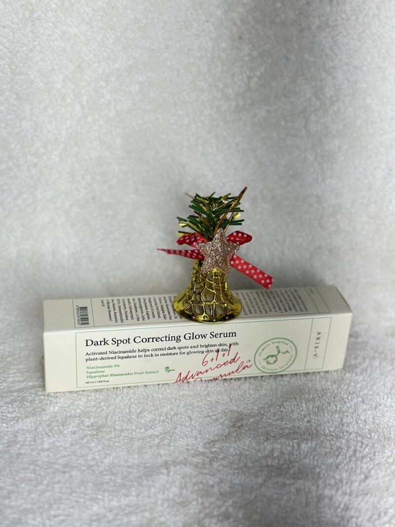A box of 'Dark Spot Correcting Glow Serum' with a small decorative bell on top, adorned with a miniature Christmas tree and a red polka-dot ribbon.
