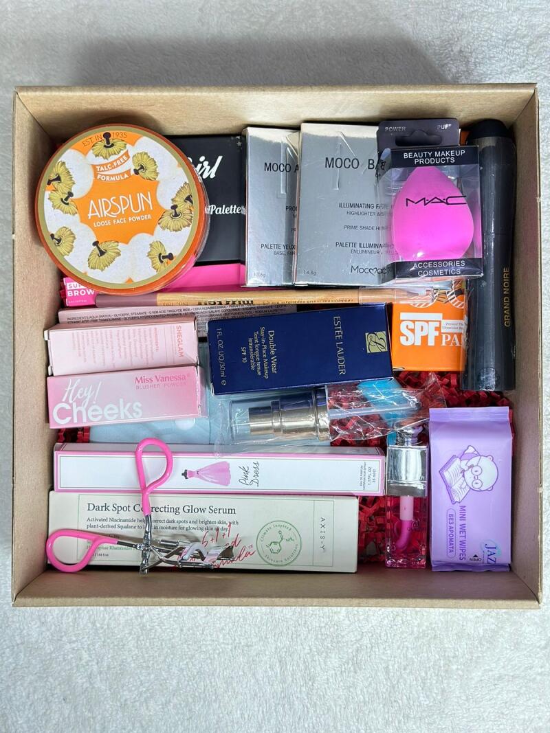 A cardboard box filled with various makeup and skincare products, including Airspun Loose Face Powder, MOCO B Illuminating Face Highlighter, MAC beauty makeup products, Estee Lauder Double Wear Stay-in-Place Makeup SPF 10, and Axis-Y Dark Spot Correcting Glow Serum.